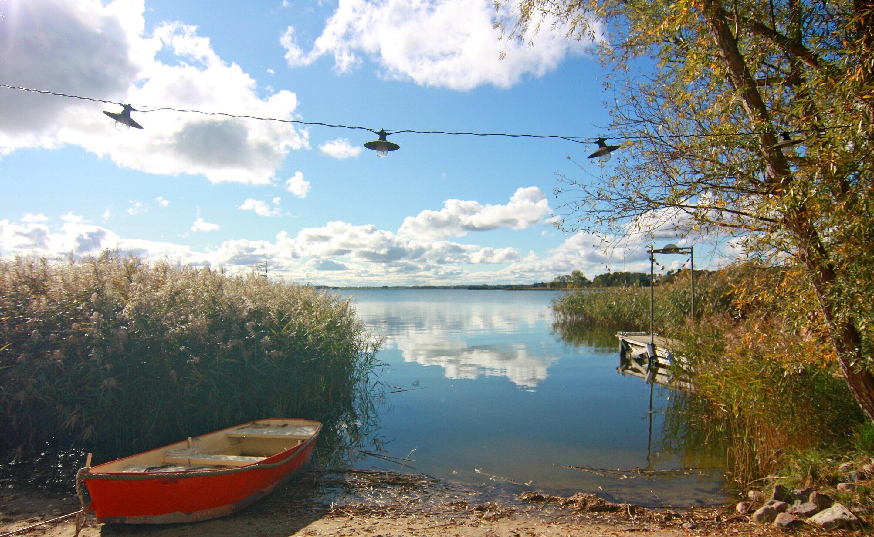 En rolig sø med en rød båd, der ligger til anker ved bredden, omgivet af vegetation og en klar himmel.