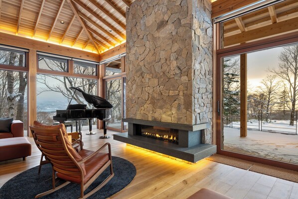 A modern interior with a stone fireplace, a grand piano, and large windows offering a view of the outdoors.