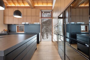 A modern kitchen with wooden cabinets, a large island, and a view of trees through a glass door.