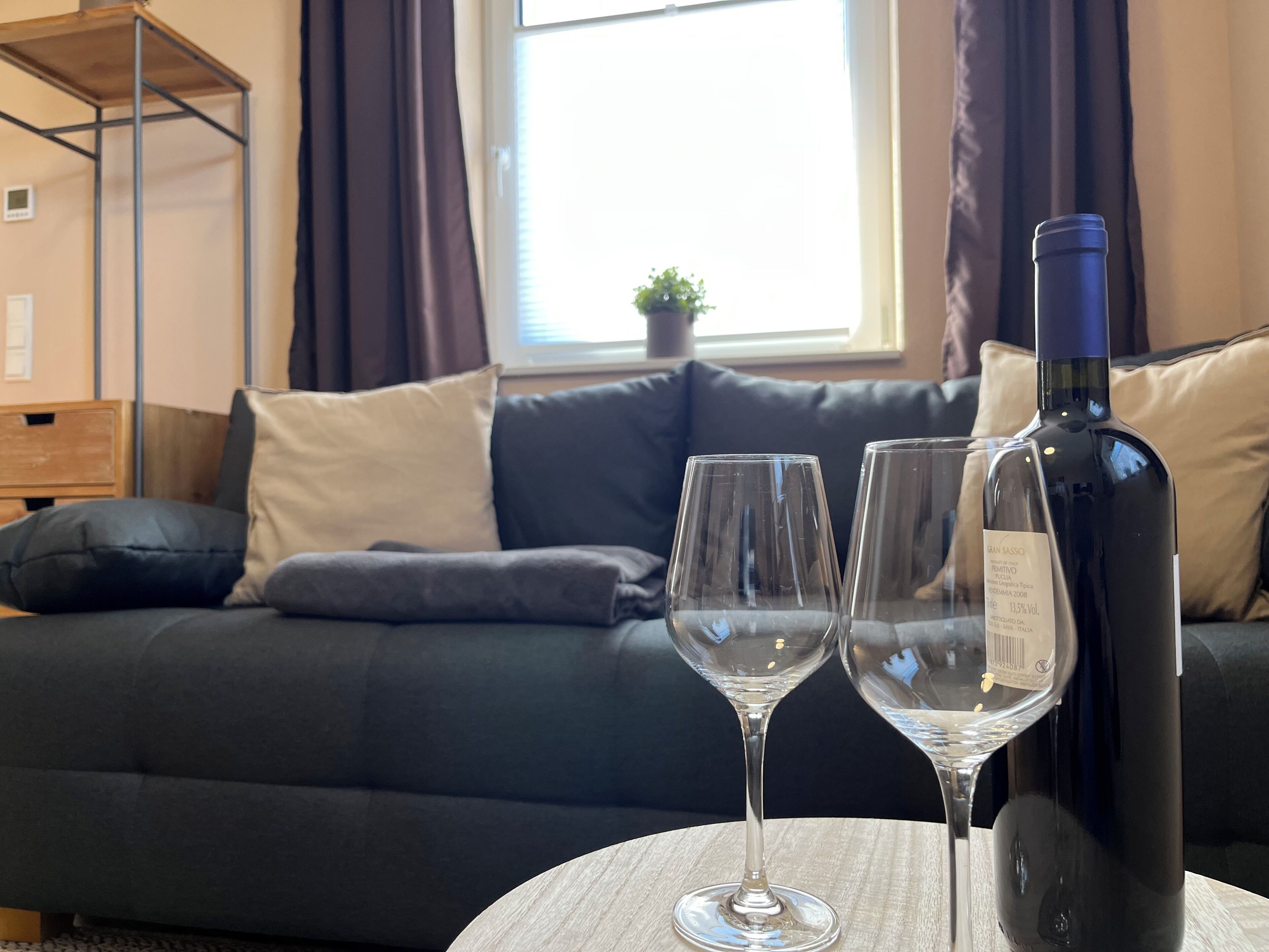 Ein Zimmer mit einem dunkelgrauen Sofa, zwei leeren Weingläsern und einer Weinflasche auf einem runden Tisch.