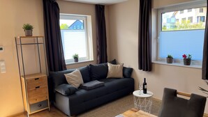 A modern living room with a sofa, a small round table with a wine glass, a chair, a tall plant stand, and two windows with curtains.