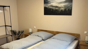 A double bed with a wooden frame, two bedside tables, a nightstand, a wall-mounted shelf, and a framed picture of a forest landscape.