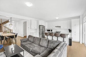 A modern living room with a sofa, a dining table, and a kitchen area with white cabinets and a stainless steel refrigerator. - 5 Bedroom Estate | Sleeps17 | 65"TV | Free Parking (Kitchener)