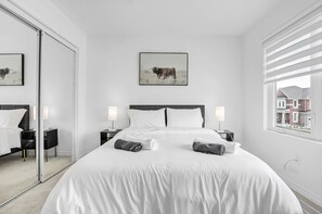 A modern hotel room with a large bed, two bedside tables, a mirror, and a window with a view of buildings. - 5 Bedroom Estate | Sleeps17 | 65"TV | Free Parking (Kitchener)