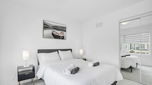 Une chambre d’hôtel moderne avec un grand lit, des tables de chevet, une petite table d’appoint, un tableau accroché au mur et une porte-fenêtre coulissante donnant sur un balcon.