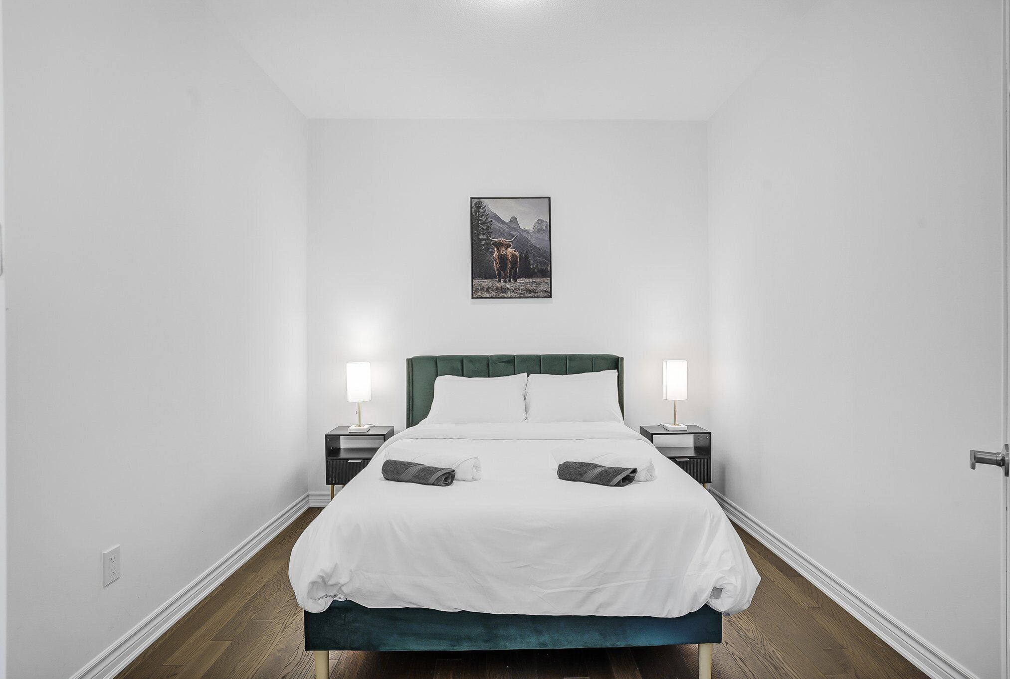 Quarto de hotel com cama, paredes brancas, cabeceira verde, duas mesas de cabeceira com abajures e um quadro na parede.