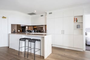 A modern kitchen with a central island, bar stools, white cabinetry, and a built-in bookshelf. - Sydney Harbour Views with Pool and Gym (Milsons Point)