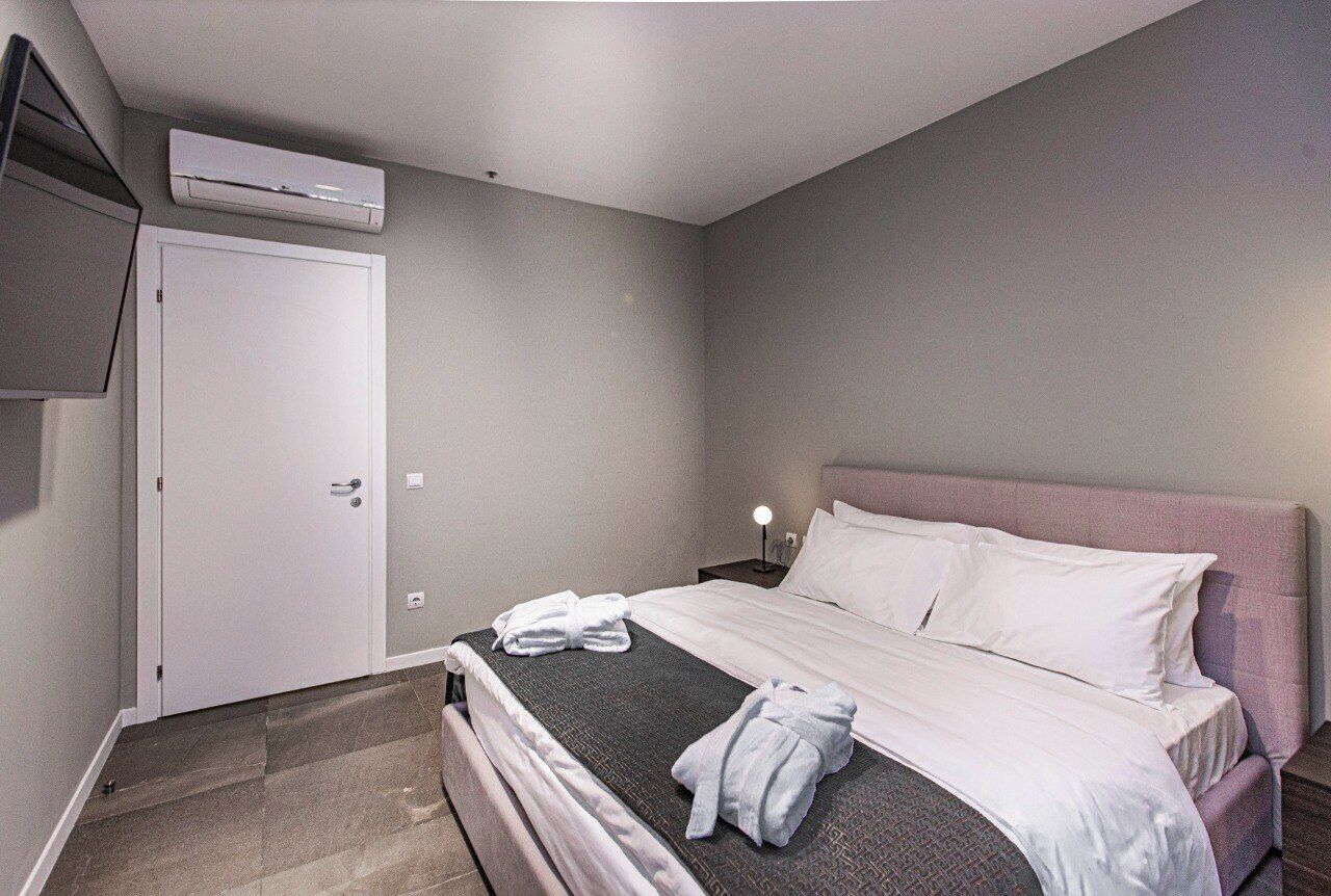 A hotel room with a bed, a bedside table with a lamp, an air conditioning unit, and a door.