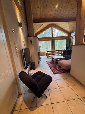 L’intérieur chaleureux d’un chalet avec un plafond en bois, un téléviseur, une cheminée, un coin repas et une grande fenêtre donnant sur les arbres.
