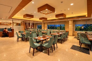 A restaurant with multiple dining tables and chairs, a reception desk, and a view of greenery outside. - Royal Aravali Udaipur (Udaipur)