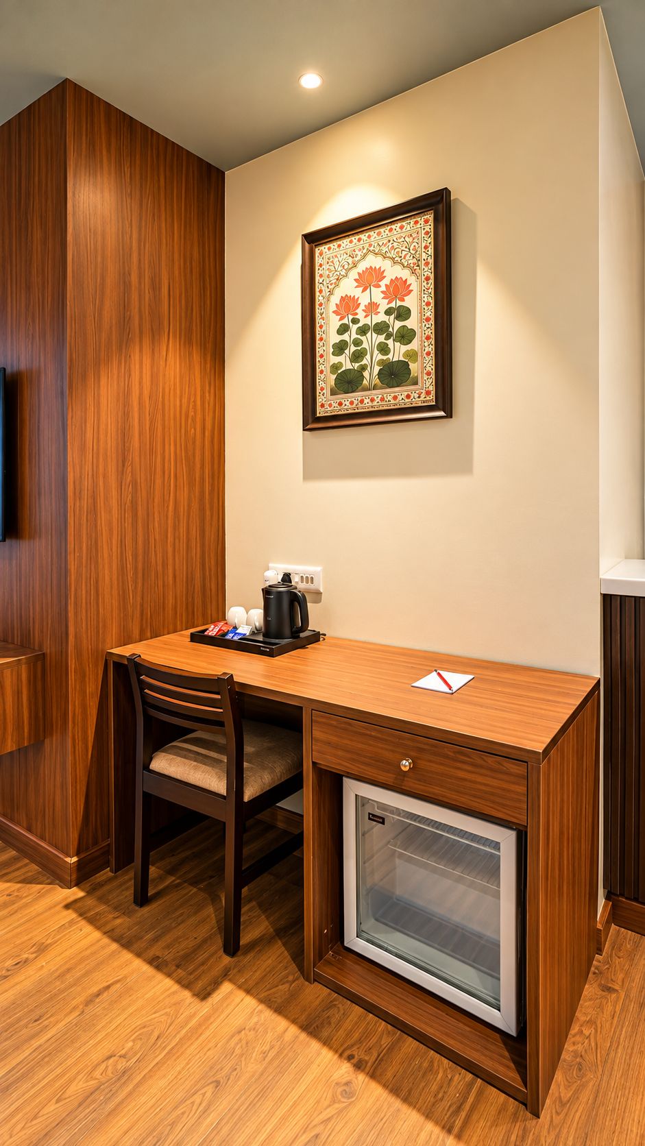 A compact office space with a wooden desk, a chair, a small refrigerator, a coffee maker, and a framed botanical illustration on the wall.