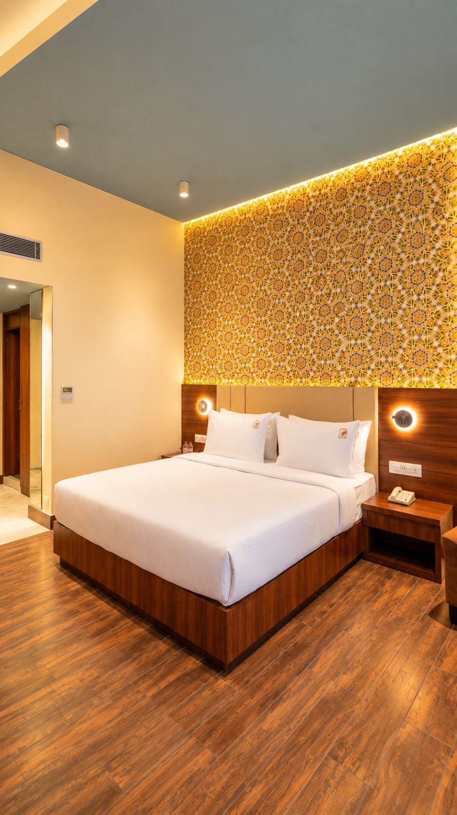 A hotel room with a large bed, wooden flooring, a patterned wall, and bedside lamps.
