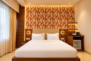 A hotel room with a large bed, a wooden headboard, a wall with a geometric pattern, bedside lamps, a nightstand, a coffee maker, and a mini fridge. - Royal Aravali Udaipur (Udaipur)