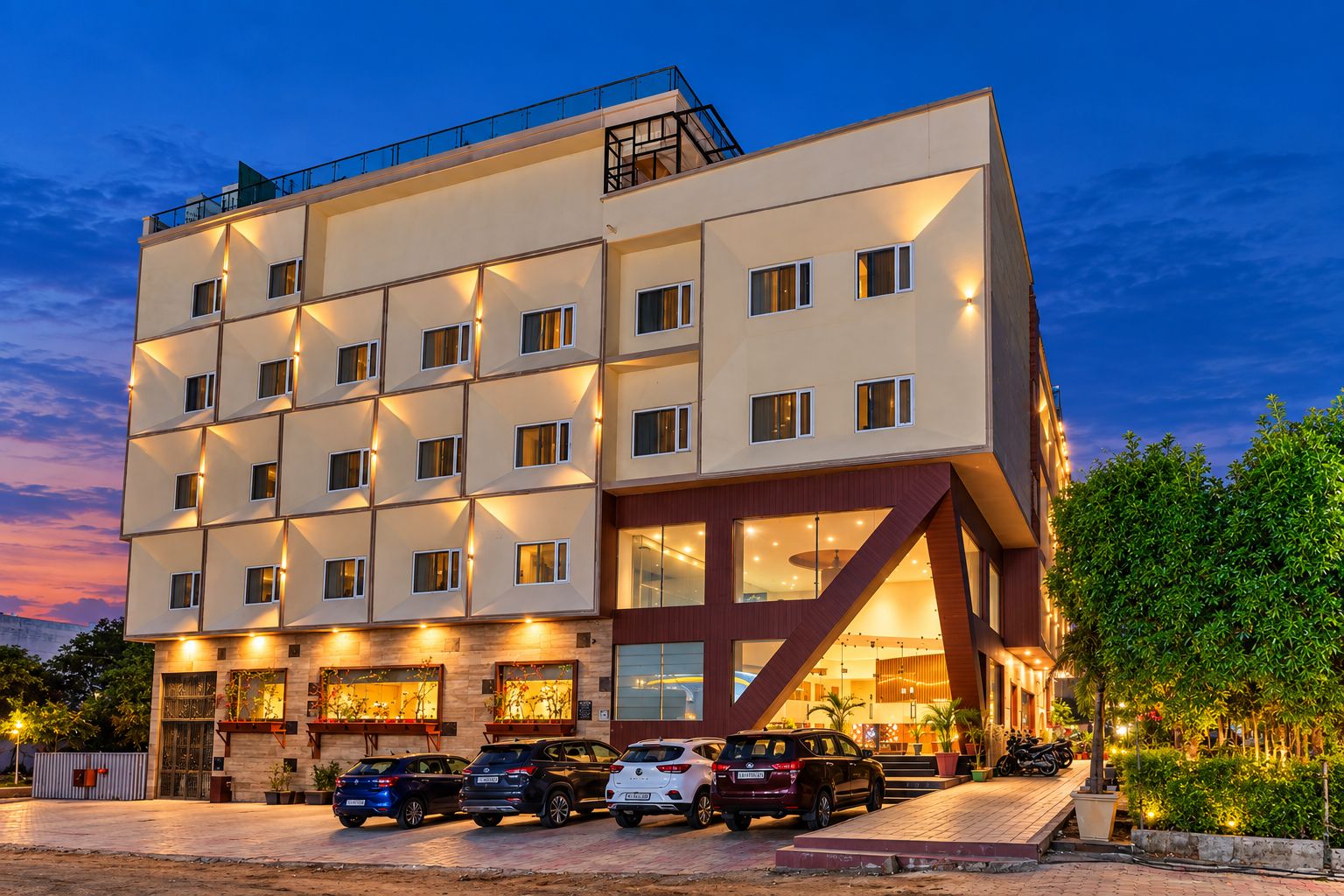 A multi-story hotel with a modern design, featuring a parking area with several cars and a well-maintained garden.