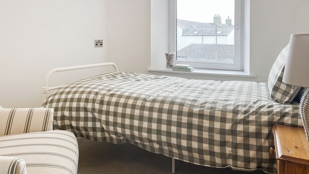 A bedroom with a bed, a bedside table, a chair, a window with a view of buildings, and a power outlet.