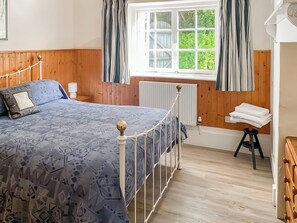 Ein Schlafzimmer mit Bett, Holzvertäfelung, einem Fenster mit Vorhängen, einem Heizkörper und einem kleinen Hocker mit Handtüchern.