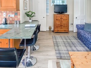 A modern kitchen with a marble countertop, a dining area with chairs, a wooden cabinet with a TV, and a blue sofa.