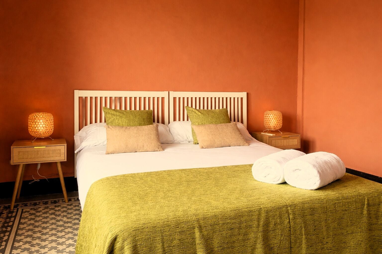 A hotel room with a bed, two bedside tables, a lamp, and a rolled towel on the bed.