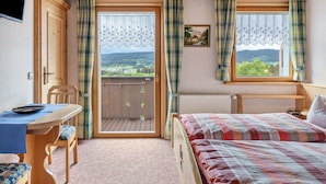 Una habitación con cama, una mesa y una silla, una ventana con cortinas, un radiador y un cuadro en la pared.