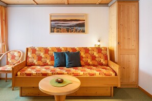 A wooden sofa with patterned cushions, a small round table, a chair, and a framed picture on the wall.
