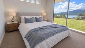 A bedroom with a large bed, two bedside tables, a view of a lake and mountains, and a window with blinds. - Kindred Whitsundays (5brm) (Woodwark)
