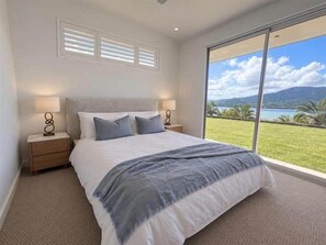 A bedroom with a large bed, two bedside tables, a view of a lake and mountains, and a window with blinds. - Kindred Whitsundays (5brm) (Woodwark)