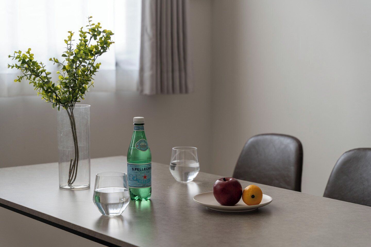 A table with a vase, a bottle, glasses, and fruit.