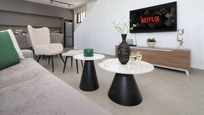 Una sala de estar moderna con un sofá, dos mesas con forma de cono negro, un jarrón con flores, una copa de vino, un aparador de madera y un televisor que muestra el logotipo de Netflix.