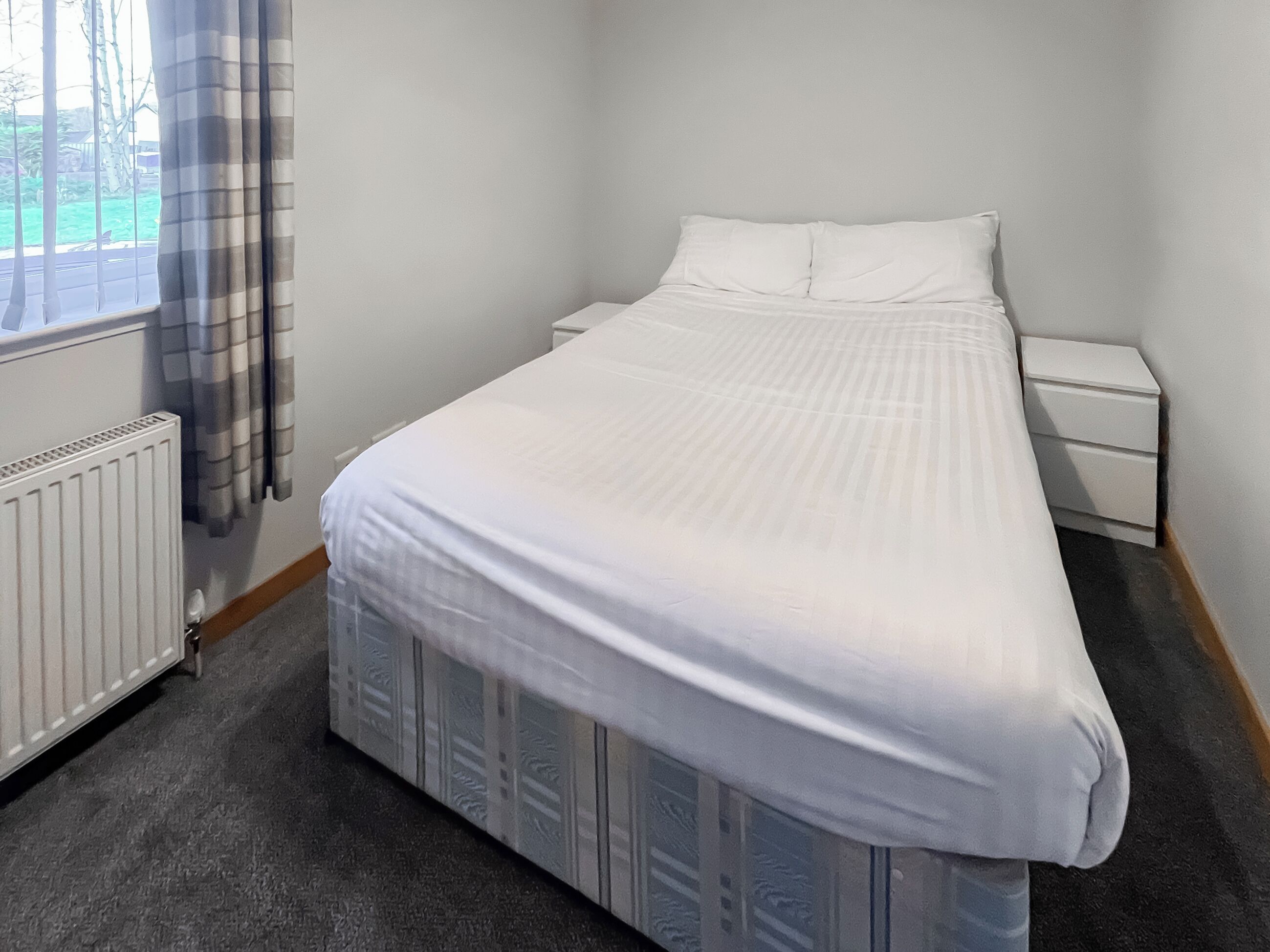 A bedroom with a bed, a nightstand, a drawer unit, a radiator, and a window with curtains.
