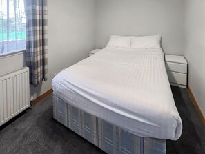 Une chambre à coucher comprenant un lit, une table de chevet, une commode, un radiateur et une fenêtre avec des rideaux.