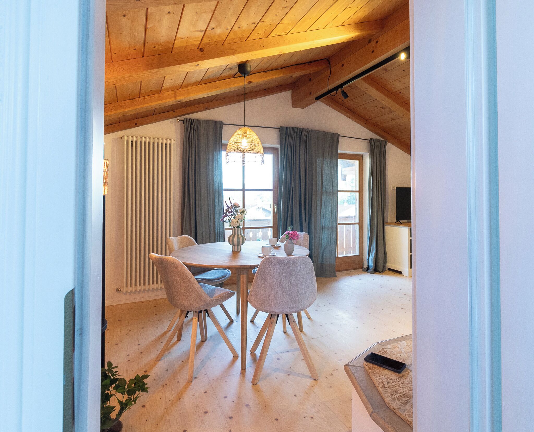 A dining area with a wooden table and chairs, a hanging light fixture, and a window with curtains.