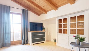 A room with a wooden ceiling, a television on a wooden stand, a cabinet with glass doors, and a small table with a vase.