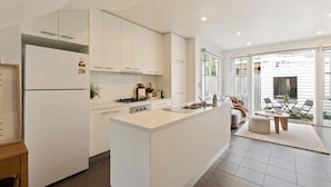 A modern kitchen with white cabinetry, a central island, and a dining area with a table and chairs. - Galatea Residence | Two Minutes to Beach and Town (Williamstown)