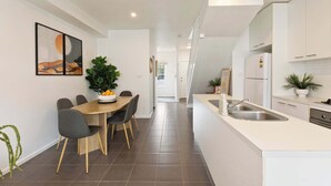 A modern kitchen with white cabinetry, a central island with a sink, and a dining area with a table and chairs. - Galatea Residence | Two Minutes to Beach and Town (Williamstown)