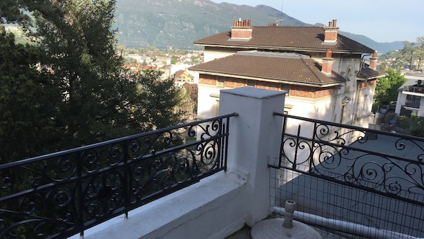 Un balcon orné d’une balustrade en fer forgé, une table ronde et une vue sur une ville, avec des bâtiments et des montagnes en arrière-plan.