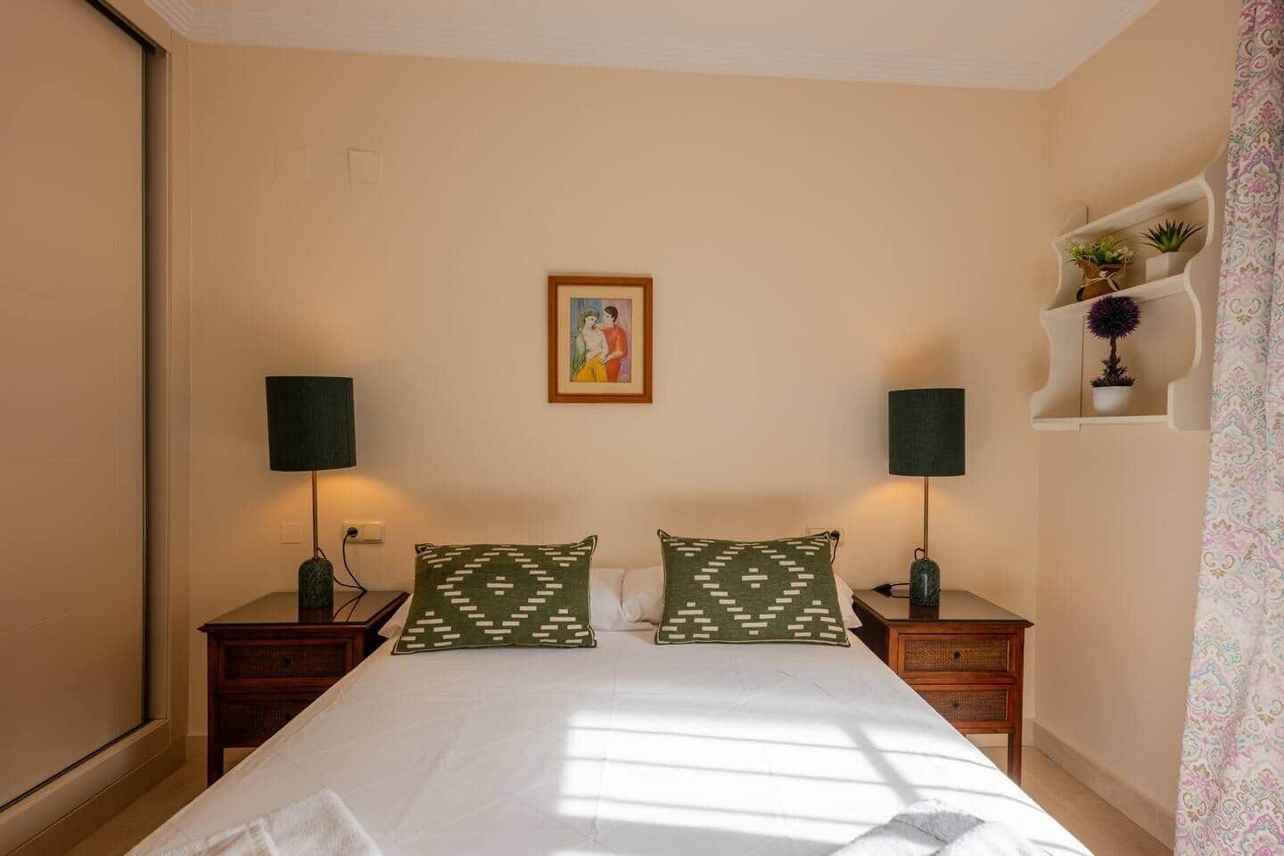 A bedroom with a bed, two nightstands, two lamps, a wall-mounted shelf with plants, and a painting.