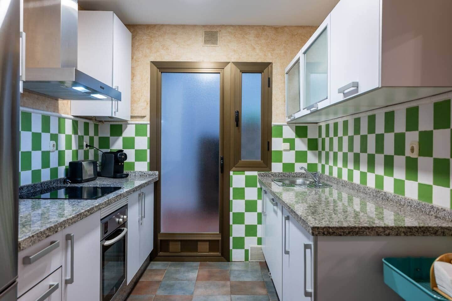 A compact kitchen with checkered green and white tiles, stainless steel appliances, and a central door.