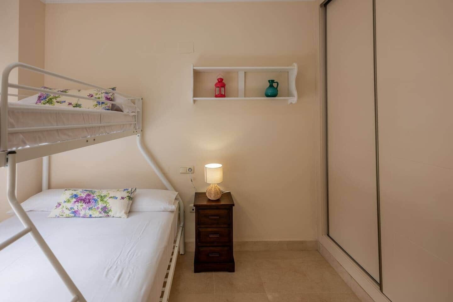 Una cama litera con un cojín floral, una lámpara de noche y una pequeña estantería con adornos.