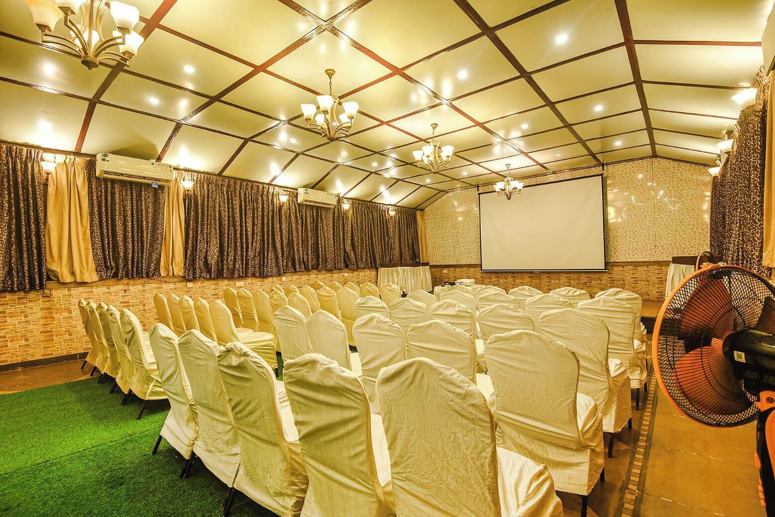Un salón de conferencias con hileras de sillas cubiertas de blanco, una pantalla de proyector y un ventilador.