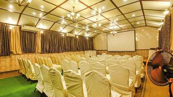 Un salón de conferencias con hileras de sillas cubiertas de blanco, una pantalla de proyector y un ventilador.