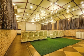 A conference room with a green carpet, white chairs, a projector screen, and a brick wall. - Hotel Shakti Palace (Ponda)