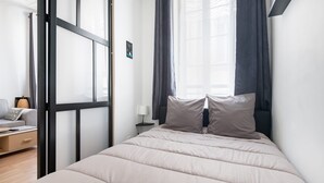A minimalist bedroom with a bed, a nightstand, a lamp, and a window with curtains. - Bohemian Studio Chic & Bright - Heart of Marseille (Marseille)