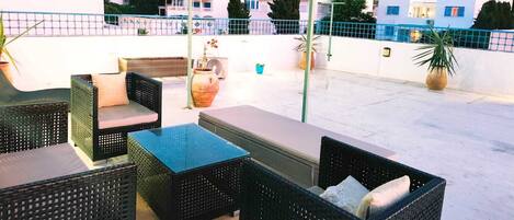 An outdoor patio with wicker furniture, a glass-top table, and potted plants.