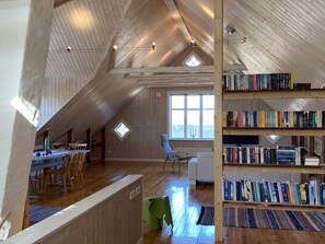 A spacious attic room with wooden beams, a large skylight, a bookshelf, and a dining area with chairs and a table.