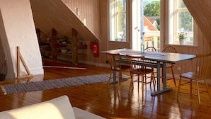 A high-ceilinged room with wooden beams, a dining table with chairs, a white sofa, and large windows with a view of houses outside. - Öland Idyll loft apartment close to the sea - swimming, surfing, kiting, hiking, cycling! (Mörbylanga)