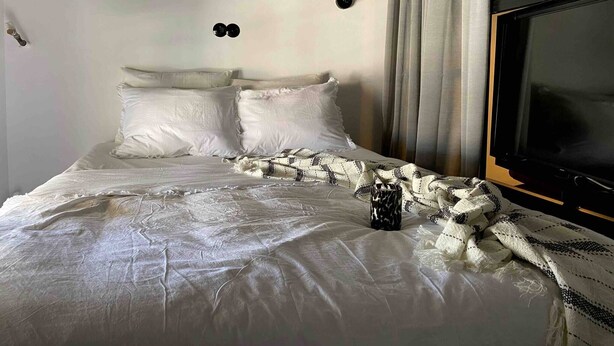 Ein Bett mit weißer Bettwäsche, zwei schwarze Wandlampen, ein Fernseher und ein Fenster mit Vorhängen.
