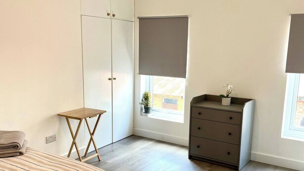 Un dormitorio con cama, cómoda, mesita, ventana con persiana e iluminación.