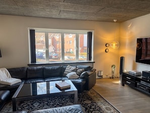 A living room with a black leather sofa, a glass coffee table, a large window with a view of buildings and parked cars, a wall-mounted TV, and a modern light fixture. - Garden Suite - 1BR - Esbjerg Island (Esbjerg)