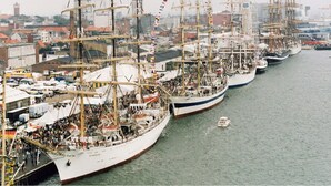 A harbor with multiple tall ships docked in a row, surrounded by buildings and a small boat in the water. - Garden Suite - 1BR - Esbjerg Island (Esbjerg)
