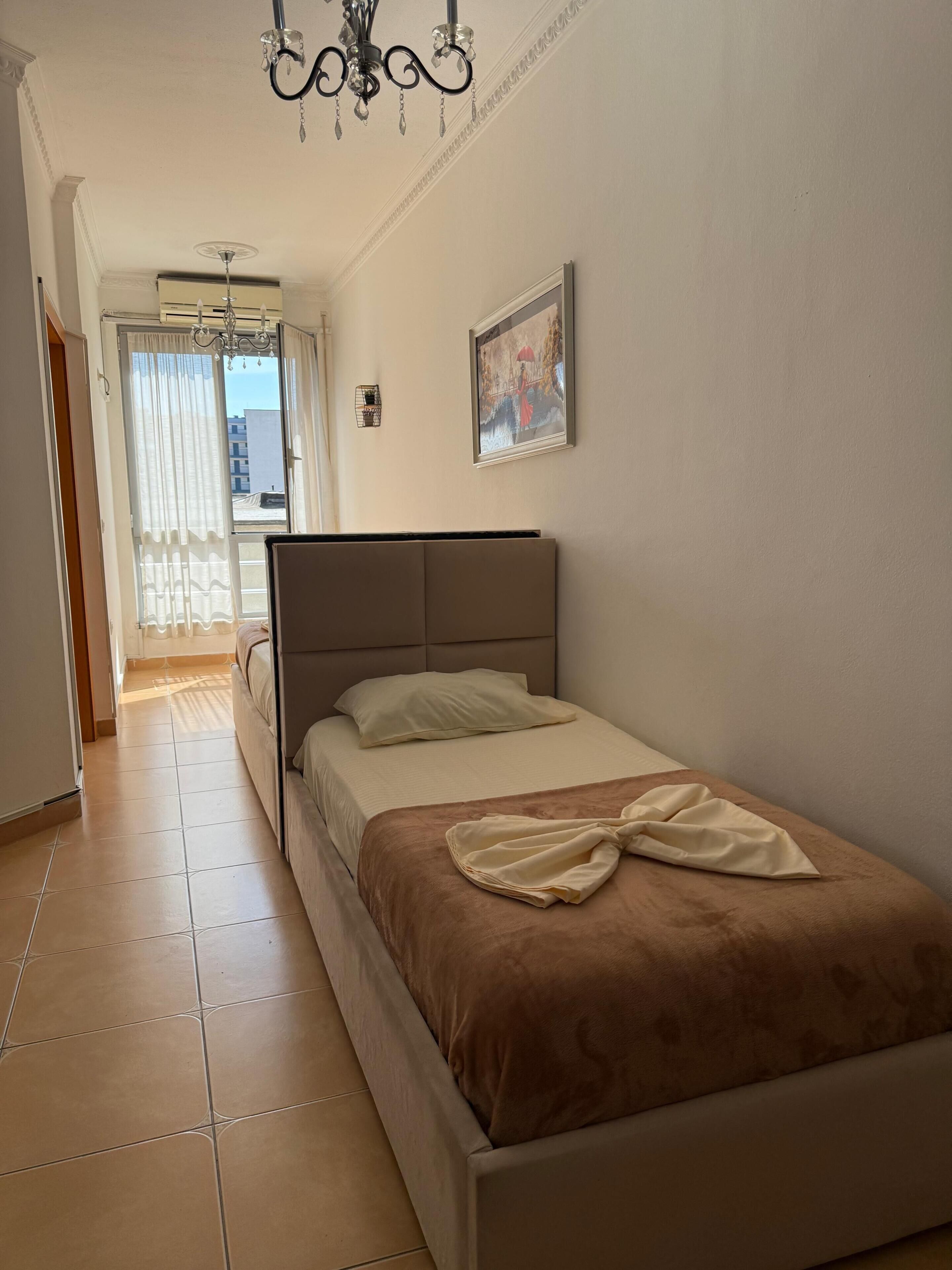 Een slaapkamer met een bed, een kroonluchter, een schilderij en uitzicht door een raam.